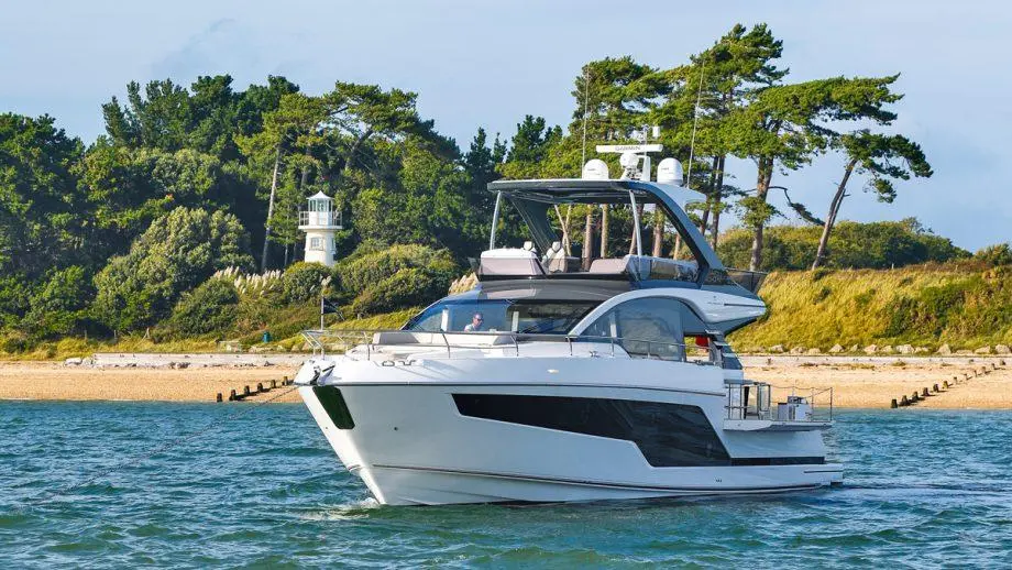 A modern yacht cruises through clear water near a sandy beach and lighthouse, surrounded by lush greenery and trees.